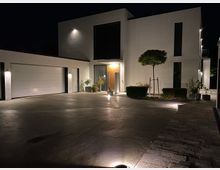 Moderne Wohnhausfassade bei Nacht, mit weißem Putz und klaren Linien. Zu sehen sind eine große Garagenfront, ein beleuchteter Eingangsbereich mit Pflanzen, ein kleiner Baum und gepflasterte sowie betonierte Flächen im Vordergrund.