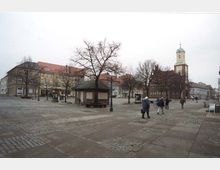 Weitläufiger Platz in einer Stadt mit gepflastertem Boden, von Bäumen und historischen Gebäuden umgeben, darunter ein Kirchturm mit einer grünen Kuppel. Einige Personen gehen über den Platz, und ein kleiner hölzerner Pavillon steht in der Mitte.