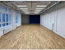 Ein leerer, rechteckiger Raum mit hellem Parkettboden, weißen Wänden und einer blauen Akzentwand im Hintergrund. An der Decke sind Rohrleitungen und Kabeltrassen sichtbar, an den Seiten befinden sich großflächige Fenster mit Heizkörpern darunter.