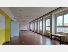 Langer, leerer Innenraum eines älteren Gebäudes mit großen Fensterreihen entlang der rechten Seite, Heizkörpern darunter und braunem Bodenbelag. Die Decke zeigt abblätternde Farbe, und am Ende des Raums stehen offene Türen sowie ein einzelner Tisch nahe den Fenstern.