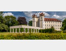 Historisches Gebäude mit heller Fassade und zwei markanten Türmen, daneben ein niedriges Nebengebäude mit großen verglasten Bögen, umgeben von gepflegten Grünflächen und hohen Bäumen.