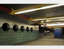 U-Bahn-Station mit modernem Design, gekennzeichnet durch grüne Fliesenwände mit runden Öffnungen, gelb-roten Rohren an der Decke und beleuchteten Schildern, darunter der Schriftzug „Rathaus Steglitz“. Der Bahnsteig ist breit und dunkel gehalten, mit elektrischer Beleuchtung.