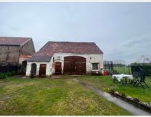 Ein rustikales Gebäude mit weiß gestrichenen, teils abgeblätterten Backsteinwänden und einem roten Ziegeldach steht in einem Garten. Im Vordergrund befindet sich eine Rasenfläche mit Gartenmöbeln und Blumenbeeten, während im Hintergrund Zaunelemente und Felder sichtbar sind.