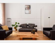 Wohnzimmer mit modernen Möbeln, darunter ein schwarzes Ledersofa, zwei schwarze Ledersessel und ein Holztisch in der Mitte. Weiße Wände mit dekorativen Paneelen, ein abstraktes Kunstwerk, eine Topfpflanze und eine minimalistische Lampe runden den Raum ab.