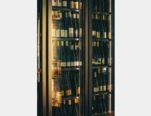 Gläserner Weinkühlschrank mit Metallrahmen und mehreren Regalböden, dicht bestückt mit Wein- und Sektflaschen. Warmes Licht und Spiegelungen in den Glastüren lassen ein Innenraum‑Ambiente wie in einer Bar oder einem Restaurant erkennen.