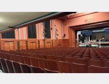Großer Theatersaal mit Sitzreihen aus Holz, einer Bühne im Hintergrund und einer Wandverkleidung in warmen Holz- und rosanen Tönen. Scheinwerfer und Bühnenelemente sind auf der Bühne sichtbar.