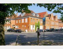 Straßenszene in einem historischen Viertel mit roten Backsteingebäuden im klassischen Stil, weißen Fenstern und Satteldächern. Im Vordergrund befinden sich geparkte Autos, Verkehrsschilder und ein öffentlicher Telefonautomat, umgeben von Bäumen.