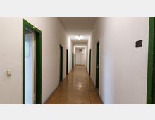 Langer, schlichter Flur mit weiß gestrichenen Wänden und mehreren Türen mit grünen Rahmen auf beiden Seiten; der Boden ist aus gelblich-braunem Linoleum, und die Beleuchtung erfolgt durch einfache Deckenlampen. Am Ende des Flurs ist eine Notausgangstür mit einem beleuchteten Exit-Schild zu sehen.