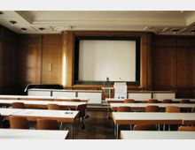 Ein Konferenzraum mit Holzvertäfelung, einer weißen Projektionsleinwand vor einer erhöhten Bühne und mehreren Reihen von Tischen und Stühlen. Die Decke ist mit dezenten Leuchten versehen und der Boden besteht aus Parkett.