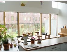 Heller Wohnbereich mit großen Fenstern, die Blick auf einen Garten und angrenzende Backsteingebäude bieten. Im Raum befinden sich ein Esstisch mit Stühlen, Pflanzen sowie eine Kücheninsel im Vordergrund.