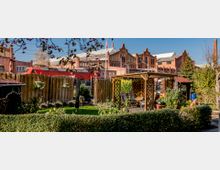 Ein kleiner Garten mit Holzpavillon, Gartenmöbeln und Blumenbeeten, umgeben von Hecken und einem Holzzaun. Im Hintergrund sind historische Backsteingebäude sowie ein vorbeifahrender roter Zug sichtbar.