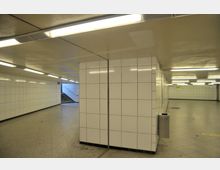 Unterirdischer Durchgang mit weißen gefliesten Wänden und einem gefliesten Boden. Im Hintergrund führen Treppen nach oben, und die Decke ist mit langen Leuchtstoffröhren beleuchtet.