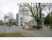 Straßenecke mit modernem weißem Mehrfamilienhaus, Bäumen ohne Laub und parkenden Autos. Im Vordergrund steht ein bemalter Altkleidercontainer, daneben Straßen- und Verkehrsschilder.