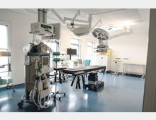 Moderner Operationssaal mit einem OP-Tisch, medizinischen Geräten wie einem Anästhesiegerät und Deckenleuchten mit integrierten Lampen. Der Raum hat helle Wände, große Fenster und einen blauen Bodenbelag.