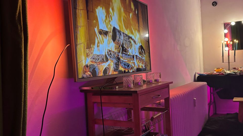 Interior of a modern living/dining room with a wall-mounted flat-screen TV showing a fireplace, lit by red and purple ambient lighting. Below the TV is a small wooden cart with shelves, a radiator along the wall, and a dining table with pendant lights in the background.