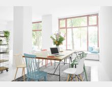 Heller Arbeits- oder Essbereich mit einem großen Holztisch, umgeben von Stühlen in verschiedenen Farben. Im Hintergrund befinden sich große Fenster mit roten Rahmen, die Tageslicht einlassen, sowie eine Fensterbank mit Kissen und Blick auf Bäume.