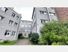 Hofbereich mit gepflastertem Weg, umgeben von grauen Wohngebäuden mit weißen Fensterrahmen; im Vordergrund stehen Büsche und ein Obstbaum mit zahlreichen grünen Äpfeln.