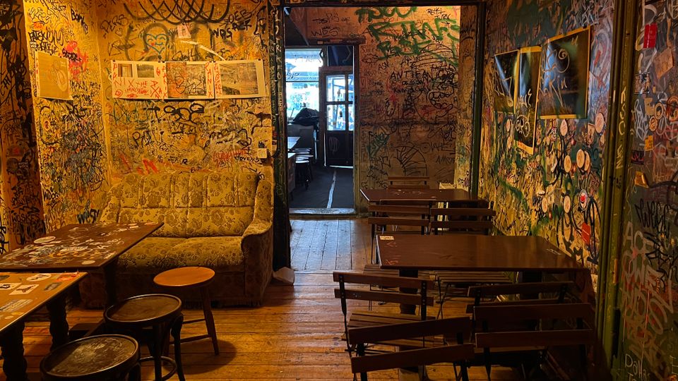 Schmaler Innenraum einer Bar oder eines Cafés mit Holzboden, warmem Deckenlicht und dicht mit Graffiti und Stickern bedeckten Wänden. Links stehen ein Sofa und kleine Tische, rechts mehrere Klapptische mit Stühlen; im Hintergrund führt ein Durchgang zu einem helleren Bereich.