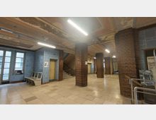 Eingangsbereich eines Gebäudes mit abgenutzten Fliesenböden, freiliegender Decke mit bröckelndem Putz und Backsteinsäulen. Die Wände sind teilweise mit hellblauen Fliesen verkleidet, eine Treppe führt nach oben und es gibt mehrere Türen sowie Fenster mit Milchglas.