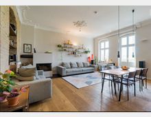 Wohnzimmer mit heller, moderner Einrichtung und Holzboden. Es gibt graue Sofas, einen Kamin, hängende Regale mit Pflanzen, große Fenster mit viel Tageslicht und einen Essbereich mit Holztisch und Stühlen.