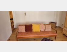 Ein schlichtes Wohnzimmer mit einer Holzbank, die ein orangefarbenes Polster und verschiedene Kissen in Gelb, Braun und gestreiftem Design hat. Der Boden besteht aus hellem Holz, und im Hintergrund ist eine weiße Tür sowie ein geflochtener Korb sichtbar.