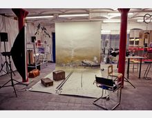 Ein Fotostudio in einem industriellen Raum mit sichtbaren Betonböden und roten Säulen. Im Vordergrund sind Requisiten wie Koffer und ein gemaltes Hintergrundbild mit einem Flugzeugmotiv, umgeben von fotografischer Ausrüstung, Stühlen und vielfältigen Dekorationselementen.