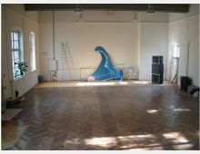 Großer, leerer Raum mit Holzboden in Fischgrätmuster, hohen Decken und hellen Wänden. An der Rückwand stehen Requisiten, darunter eine blaue Welle, eine Leiter und mehrere Lautsprecher; links befinden sich große Fenster mit Gitterrahmen.