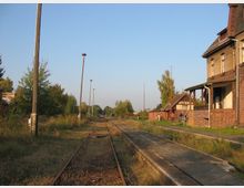 Ehemaliger ländlicher Bahnhof mit verfallenen Gleisen und Bahnsteigen, flankiert von einem Backsteingebäude rechts und Bäumen im Hintergrund. Straßenlaternen säumen die Gleise, und Reste von Vegetation sind entlang des Geländes sichtbar.