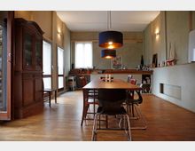 Esszimmer in einem modernen, aber rustikal eingerichteten Raum mit Holzboden. Ein langer Holztisch mit verschiedenen Stühlen steht im Zentrum, darüber hängen zwei dunkle Lampen. An der Rückwand befindet sich ein Regal mit Dekorationsobjekten und Geschirr, daneben eine Vitrine aus Holz.