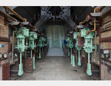 Industrieller Maschinenraum mit historischen hydraulischen Pumpen, die in zwei symmetrischen Reihen an den Wänden montiert sind; im Hintergrund eine Treppe, die zu zweiflügeligen, grünen Glastüren führt. Die Umgebung besteht aus Ziegelwänden und sichtbaren Metallrohren und Hebeln.