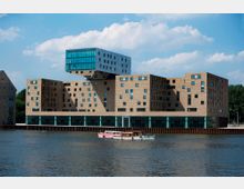 Modernes Gebäude am Wasser mit einer Fassade aus gelblichen Backsteinen und vielen unregelmäßig angeordneten Fenstern. Auffällig ist ein überhängender weiß-blauer, würfelförmiger Abschnitt im oberen Teil; im Vordergrund fließt ein Fluss mit einem kleinen Boot.