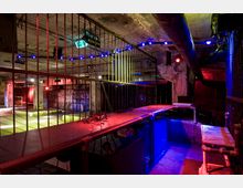 Unterirdischer Raum mit industriellem Ambiente, gekennzeichnet durch Betonwände, Rohre und Gitterabsperrungen. Farbige Beleuchtung in Blau, Rot und Gelb sorgt für eine Club-ähnliche Atmosphäre.