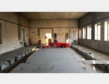 Großer, leerer Industrie- oder Lagerraum mit unverputzten Ziegelwänden, Betonboden und rechteckigen Fenstern, durch die Tageslicht einfällt. In der Mitte des Raumes stehen einige rote Möbelstücke und Gegenstände, während Teile des Bodens abgesenkt oder unvollständig sind.