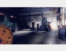 Industriehalle mit sichtbaren Maschinen und metallischen Strukturen, darunter eine große mechanische Vorrichtung in der Mitte. Die Wände sind aus Ziegeln, teils verputzt, mit hohen Fenstern, durch die schwaches Licht fällt.