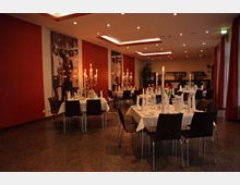 Ein festlich dekorierter Veranstaltungsraum mit weiß gedeckten Tischen, auf denen Kerzenleuchter, gefaltete Servietten und Gläser arrangiert sind. Der Raum hat rote Wände, große Wandbilder, eine dezente Beleuchtung und lange Vorhänge an den Fenstern.