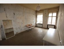 Ein leerer Raum mit abgenutzten, teils abgeblätterten Wänden, zwei großen Fenstern mit weißen Vorhängen, einem Heizungskörper darunter und einem altmodischen Lampenschirm an der Decke. Im Raum stehen zwei Stühle, ein rechteckiger Spiegel an der Wand und ein weißer Tisch.