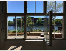 Blick aus einem offenen Fenster auf einen Fluss mit Vegetation am Ufer und Gebäuden im Hintergrund. Im Vordergrund sind ein Holztisch und Teile eines Innenraums zu sehen.