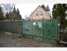 Grünes Metalltor mit Graffiti, das den Eingang zu einem Grundstück mit einem kleinen Haus im Hintergrund absperrt; das Haus hat ein Giebeldach und helle Fassaden, umgeben von Bäumen und einem Maschendrahtzaun. Der Boden vor dem Tor ist aus Kopfsteinpflaster.