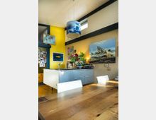 Moderne Küche mit offenem Wohnraum; zentral eine kleine Kücheninsel mit grauen Schubladen und schwarzer Arbeitsplatte. An der gelb gestrichenen Wand hängen Bilder, darüber eine Hängelampe mit Wolkenmotiv. Im Vordergrund ein Holztisch mit weißen Stühlen.