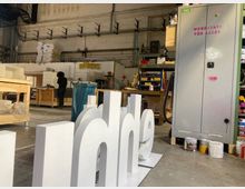 Eine Werkstatt mit hohen Wänden, Regalen und Arbeitsbereichen. Im Vordergrund stehen große, weiß gestrichene Buchstaben, während im Hintergrund eine Person an einer Werkbank arbeitet und der Schriftzug „NERDS“ an der Wand hängt.