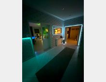 Innenansicht eines Flurs in einer Wohnung mit weiß gefliesten Böden, gemusterter Tapete und mehreren Türen zu angrenzenden Räumen. LED-Lichtleisten rahmen Tür- und Wandöffnungen, und an der Wand befinden sich ein Heizkörper sowie ein schmaler Teppichläufer auf dem Boden.