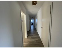 Ein langer, heller Flur in einem modernen Innenraum mit weiß gestrichenen Wänden, Holzboden und mehreren geschlossenen Türen auf beiden Seiten. Am Ende des Flurs ist ein Fenster mit Tageslicht und das entfernt sichtbare Mobiliar erkennbar.