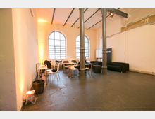 Ein heller, industrieller Raum mit hohen Decken und großen Rundbogenfenstern. Der Raum enthält moderne Sitzbereiche mit Tischen und Stühlen, schwarzen Sofas, sowie freistehende Metallpfeiler zur Unterstützung der Struktur.