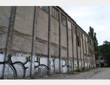 Backsteinfassade eines verlassenen Industriegebäudes mit verblassten Graffitis im unteren Bereich. Der Asphalt davor ist rissig und überwuchert, im Hintergrund stehen vereinzelte Bäume und weitere Gebäuderuinen.