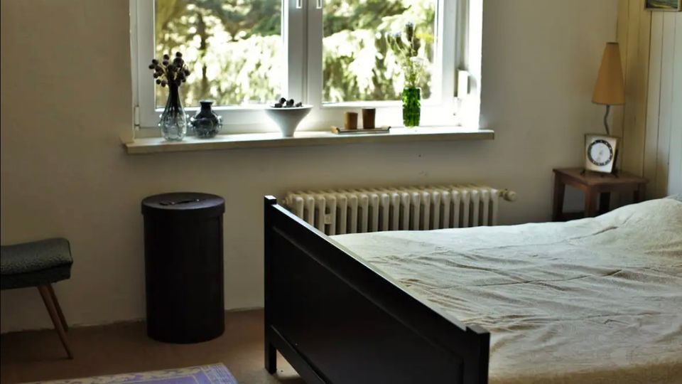 Schlafzimmer mit einem einfachen Bettgestell in dunklem Holz, einem kleinen Nachttisch mit Lampe und Wecker sowie einer Fensterbank mit Dekorationen wie Vasen und Schalen. Das Fenster bietet Ausblick auf grüne Bäume, und unter der Fensterbank befindet sich ein Heizkörper.