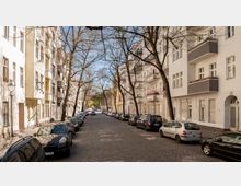 Eine ruhige, gepflasterte Wohnstraße mit mehrstöckigen Altbauten auf beiden Seiten, Balkonen und geparkten Autos entlang der Straße. Bäume mit kahlen Ästen säumen die Straße und spenden Schatten, während im Hintergrund ein Stück Grünanlage sichtbar ist.