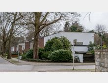 Straßenszene in einem Wohngebiet mit modernen Reihenhäusern, gekennzeichnet durch weiße und rötliche Fassaden. Im Vordergrund stehen große Bäume und grüne Büsche, während ein Straßenschild mit der Aufschrift "Dohnenstieg" sichtbar ist.