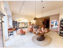 Innenraum eines modernen Cafés mit gemütlicher Einrichtung. Zu sehen sind ein großer Holztisch mit dekorativen Produkten, rosa Sessel, zwei Regalwände mit Waren, ein Kühlschrank mit Getränken und ein Durchgang zu einem weiteren Bereich, der in rosafarbenem Licht beleuchtet ist.