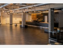 Offene Innenansicht eines modernen Büro- oder Veranstaltungsraums mit industriellem Design, sichtbar durch sichtbare Betonpfeiler, freiliegende Metallrohre und hängende Glühbirnen. Im Hintergrund befindet sich eine Theke mit Holz- und Metallelementen, flankiert von weißen Wänden und einer glatten, dunklen Bodenfläche.