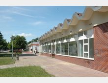 Straßenansicht eines eingeschossigen Gebäudes mit großer Fensterfront und markantem gezacktem Dach, umgeben von einem gepflasterten Gehweg und einer Grünfläche. Im Hintergrund sind Bäume, weitere Gebäude und vereinzelte Personen zu sehen.